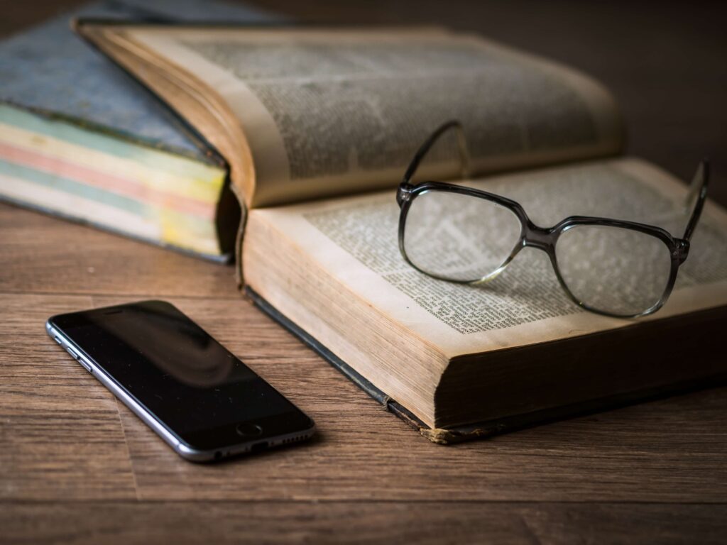 本と眼鏡、スマートフォンが並んだ机。情報を読み、調べ、考えるイメージ。